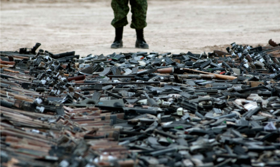 A pile of guns scattered on the ground with a cropped view of a person's lower extremities